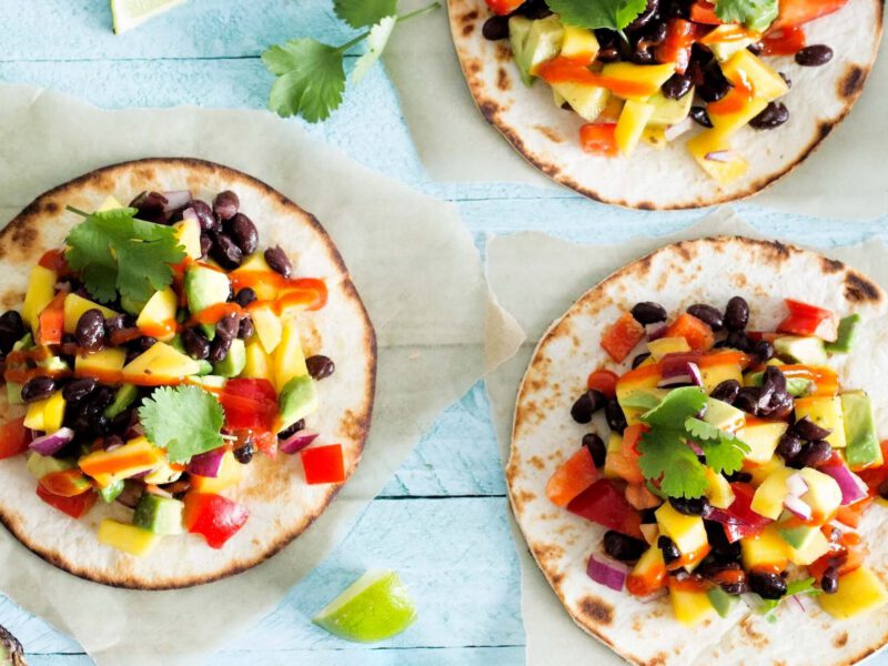 Three Mango and Black Bean Tacos, each on a small piece of parchment paper, against a pale blue wooden background, with fresh mango, avocado, and lime dotted around them