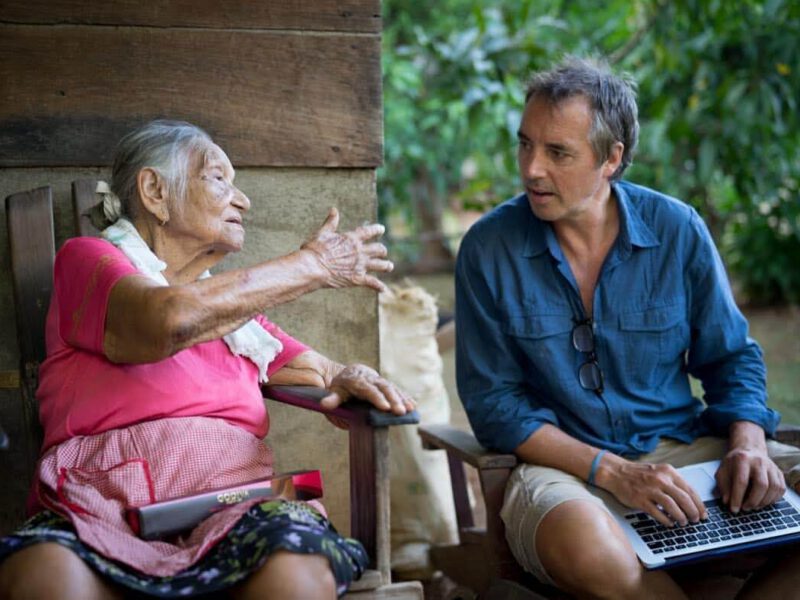 Dan Buettner sitting with a lap top on his lap talking to a very old woman