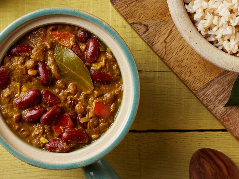 Top down view of Kidney Bean and Lentil Dal on a mustard counter next to a wooden chopping board, a bowl of brown rice, and cinnamon sticks