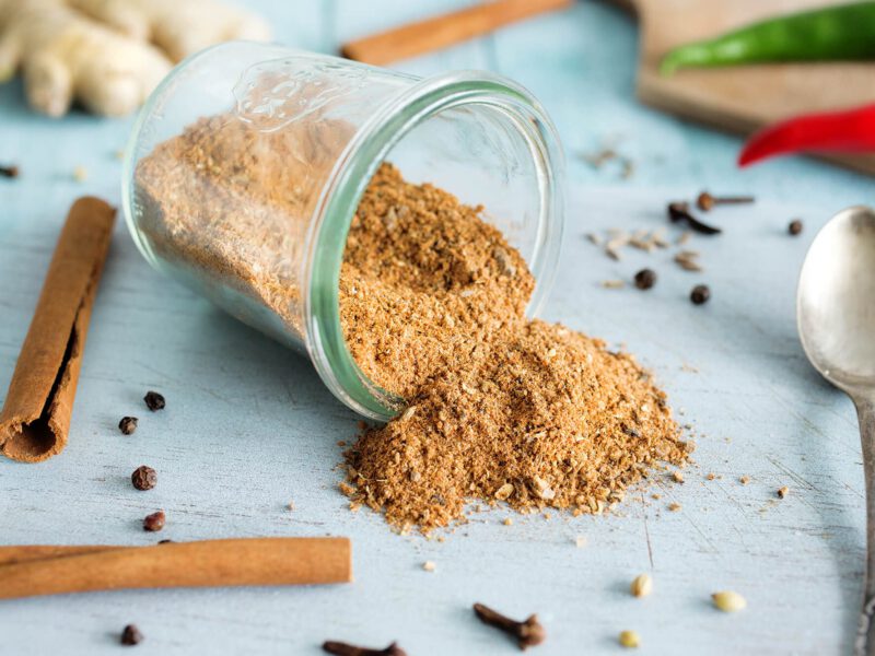 A small mason jar on it's side on a pale blue wooden counter, with a seasoning spilling out. Around the jar are cinnamon sticks, fresh ginger, cloves, and fresh chiles.