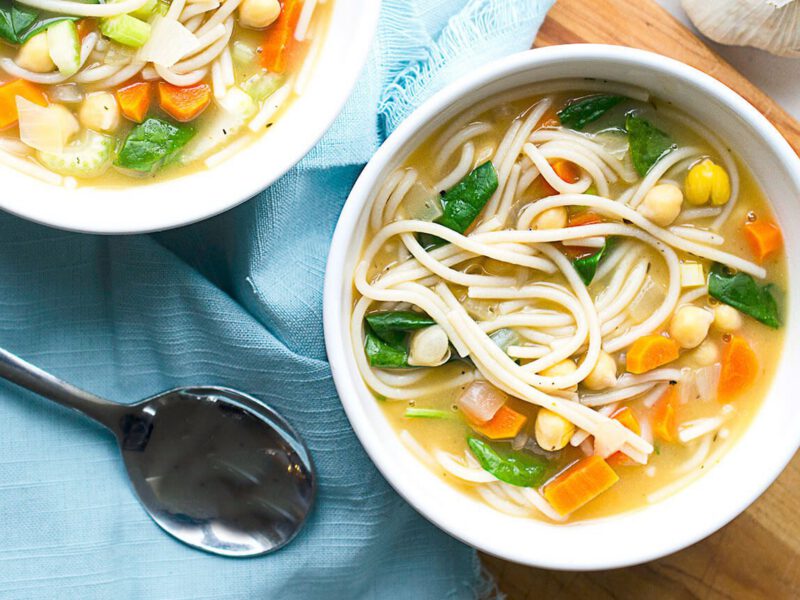Two bowls of 30-Minute Chickpea Noodle Soup with Spinach with fresh onion and garlic to the side