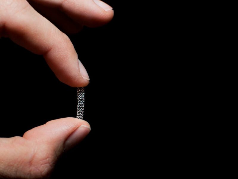 Two fingers holding up a stent, against a black background