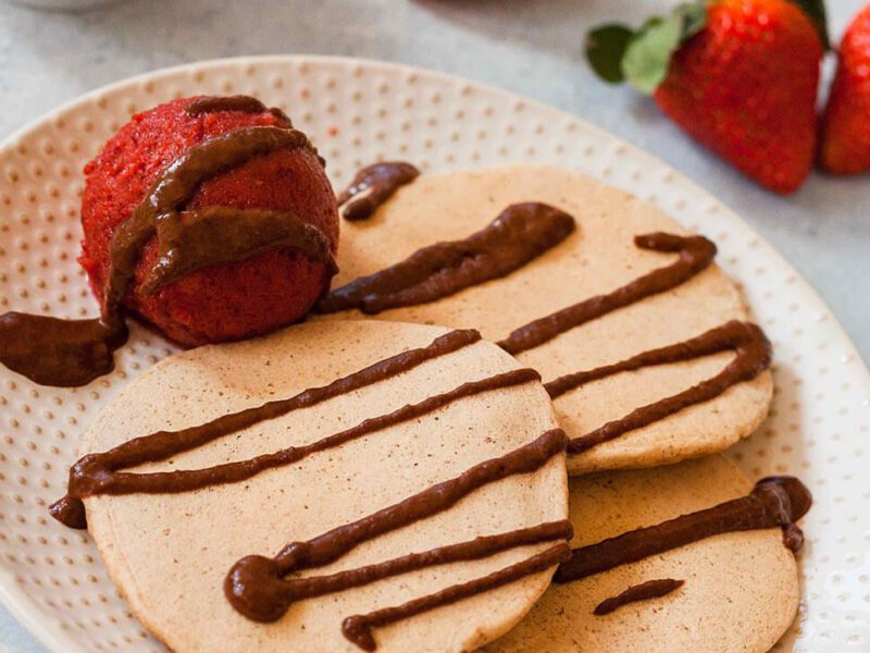 Vanilla Pancakes with a scoop of Strawberry-Cherry Ice Cream drizzled with Warm Chocolate Sauce. In the background is a bowl of chocolate sauce, a bowl of ice cream, and fresh strawberries