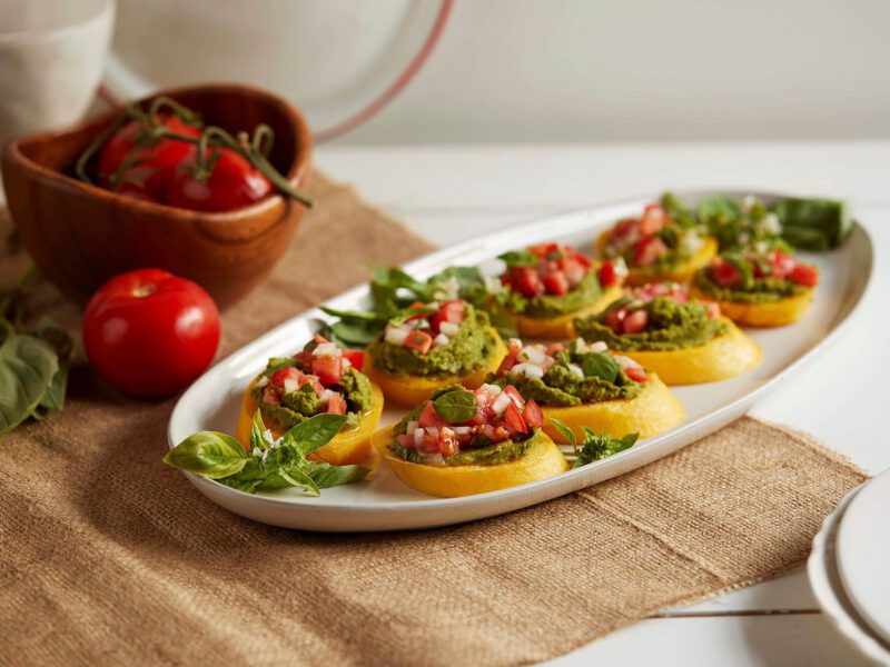 Polenta Crostini with Chickpea Pesto on a white oval serving platter on a hessian bag next to fresh basil and fresh tomatoes