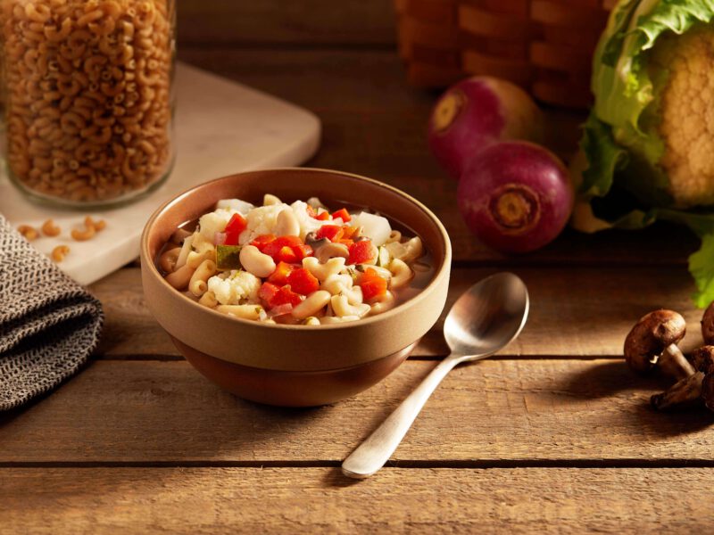 Winter White Bean Minestrone Soup in a brown ceramic bowl on a rustic wooden table top with raw vegetables and a jar of dried macaroni