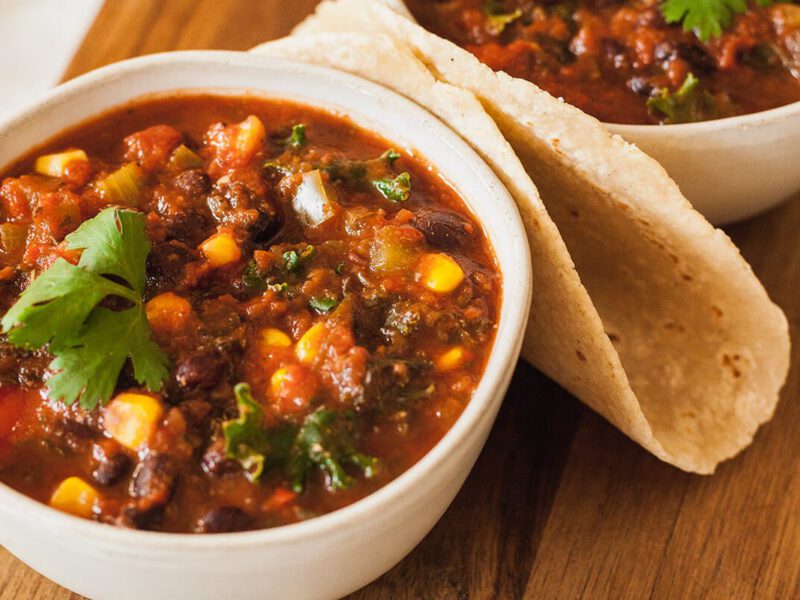Two bowls of Black Bean and Corn Chili on a wooden chopping board with a warm tortilla folded