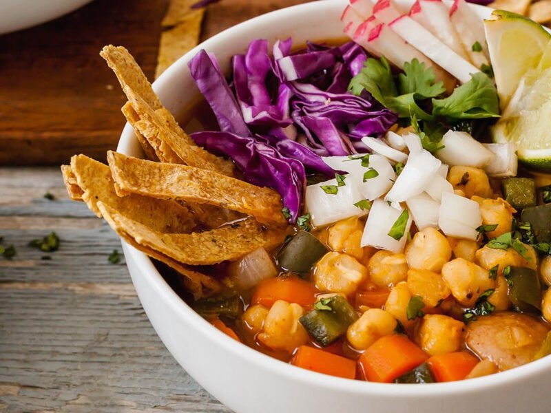 A white serving bowl Vegan Pozole garnished with lime wedges, tortilla strips, finely chopped red cabbage and onion