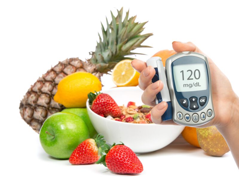 A colorful array of fruits such as lemon, pineapple, and strawberries. In the foreground, a hand holds a glucose monitor