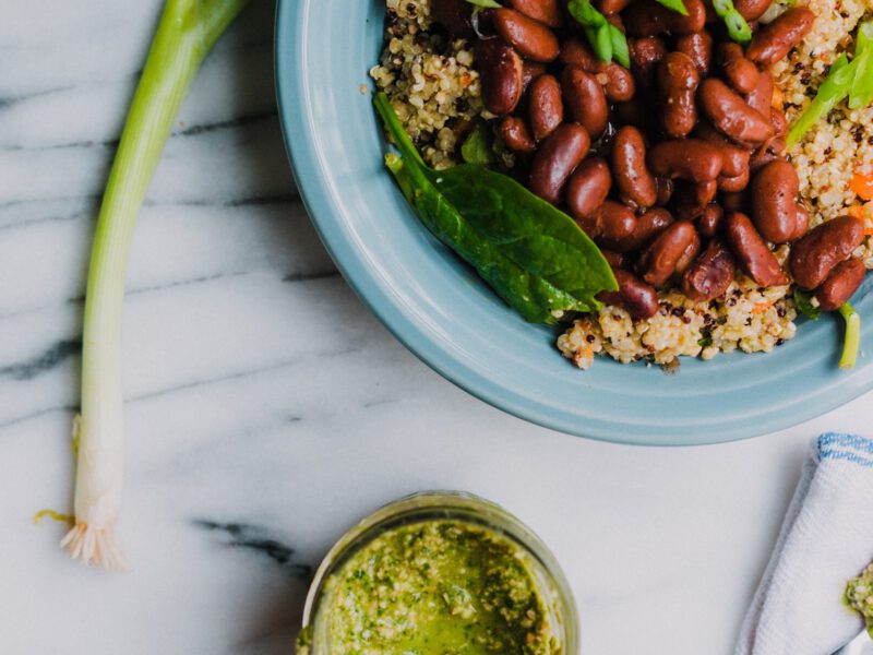 A blue bowl of quinoa, kidney beans, and greens on a marble couner next to green onions and a small bowl of green sauce