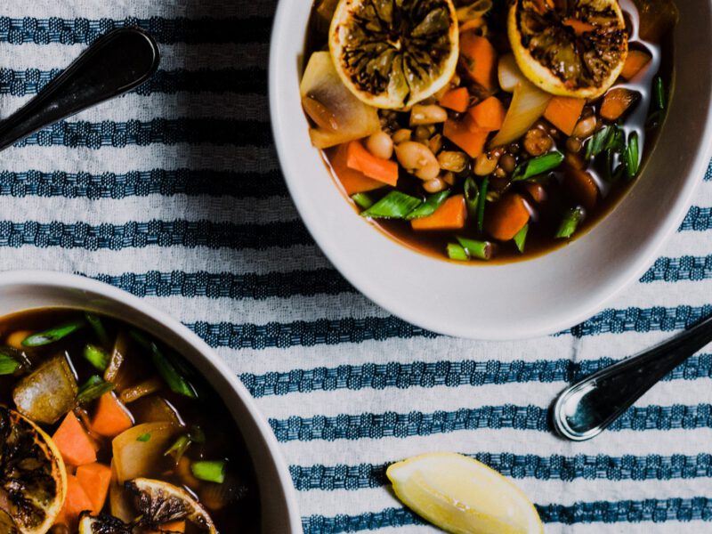 Two bowls of vegan stew on a white and blue tablecloth with two spoons and wedges of lemon