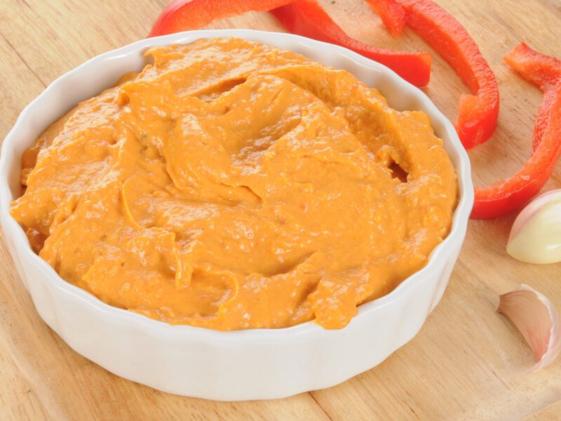 Roasted Red Pepper Tapenade Hummus in a white bowl next to fresh garlic cloves a slices of red bell pepper
