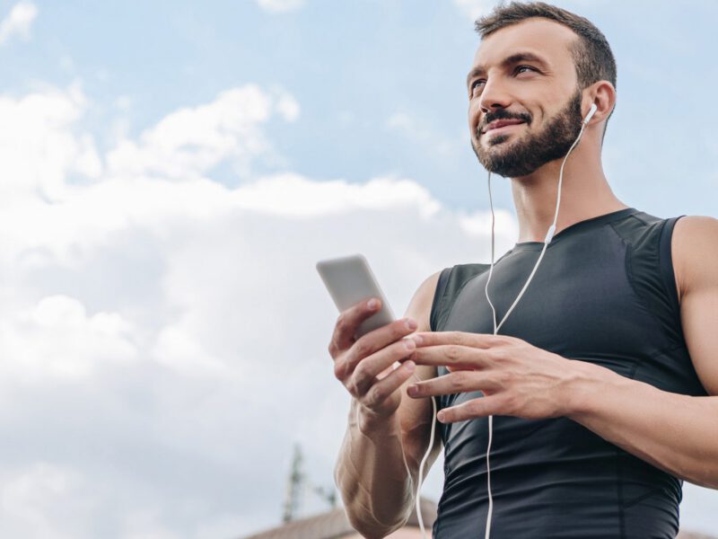 A man in a sleeveless top with the blue sky above him, holding his phone, with ear buds in his ears, listening to a plant-based podcast