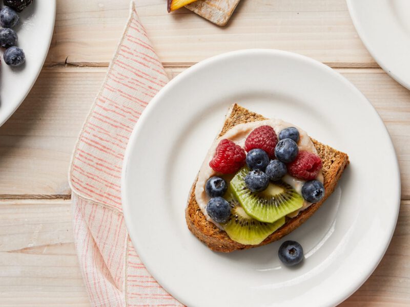 Two plates of Sweet White Beans on Toast with Berries next to a plate of fresh fruit, including kiwi slices, raspberries, and blueberries