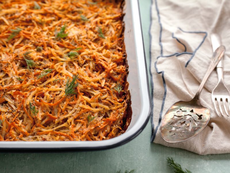 A casserole dish of Sweet Potato Kugel next to a white kitchen towel and silver serving spoons