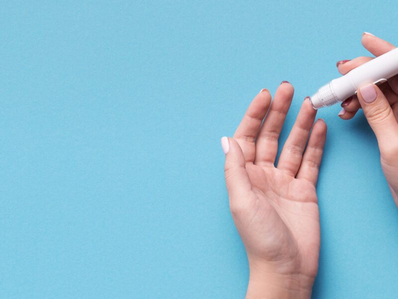Two hands shown against a blue background as the persons pricks their finger so they can test their blood sugar