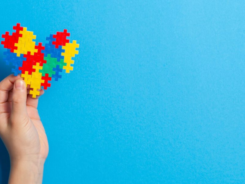 A hand holds up a heart made of colorful jigsaw pieces, against and blue background