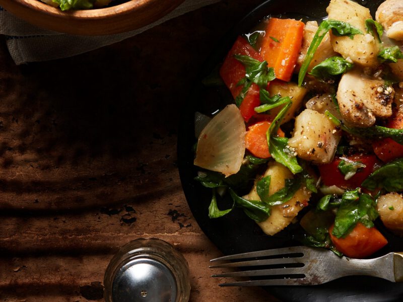 A black plate of Jerk-Seasoned Veggies with Yuca with a wooden-handled fork resting in the plate