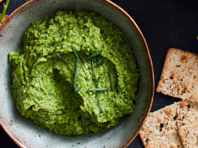 Emerald Hummus Dip with Fresh Herbs garnished with chives, with whole grain crackers around the bowl
