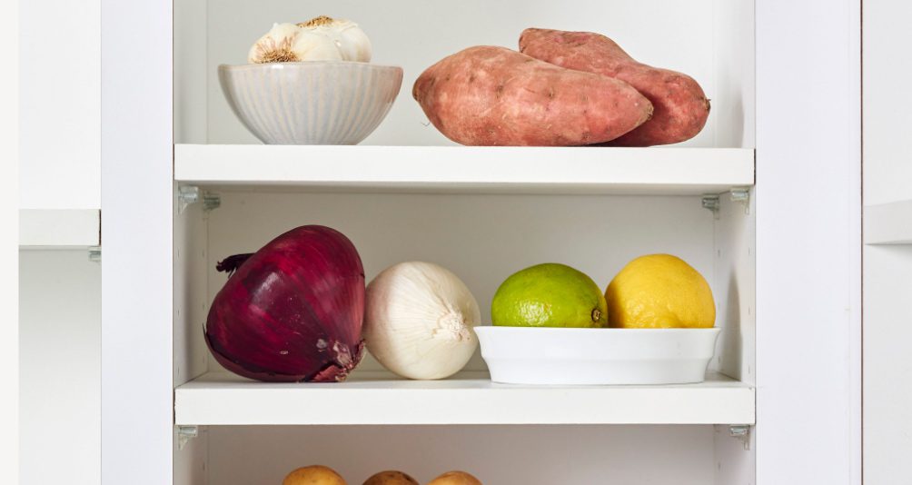 a white cabinet showing fresh pantry items such as a red onion, garlic, potatoes, lemon and lime