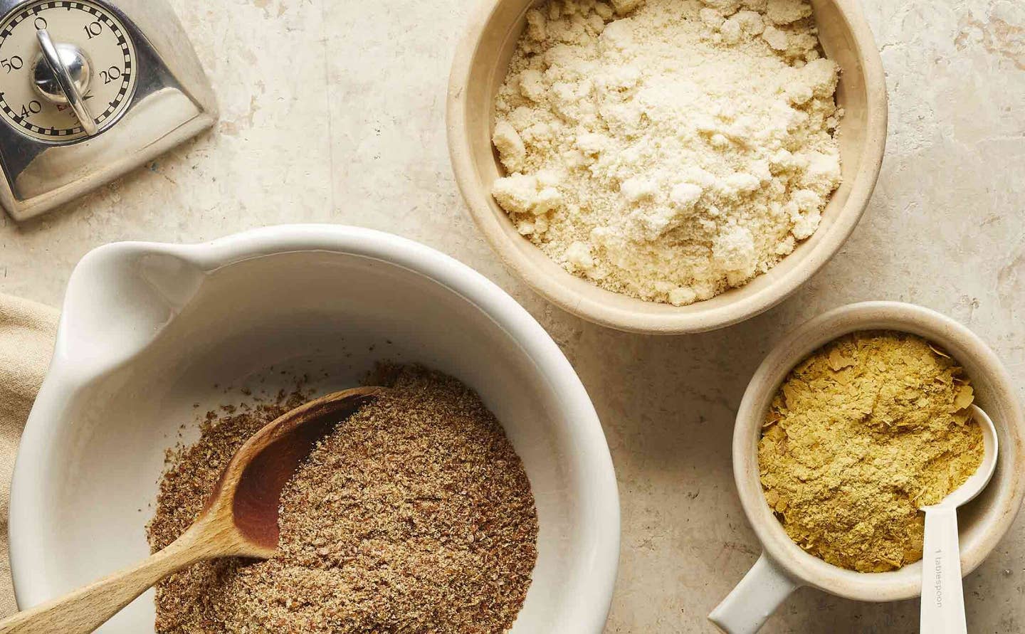 Various ingredients for gluten-free baking, including a bowl of potato starch, ground flaxseeds, and cornmeal or corn flour, with a kitchen timer off to the side