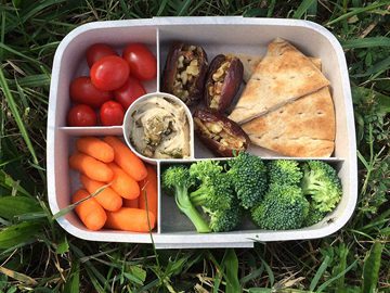 Bento box with baby carrots, broccoli, sliced pita, dates, and cherry tomatoes organized into compartments