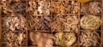 An array of whole wheat pasta shapes in a wooden crate, with compartments dividing each shape, including penne, orecchiette, spaghetti, and rotini