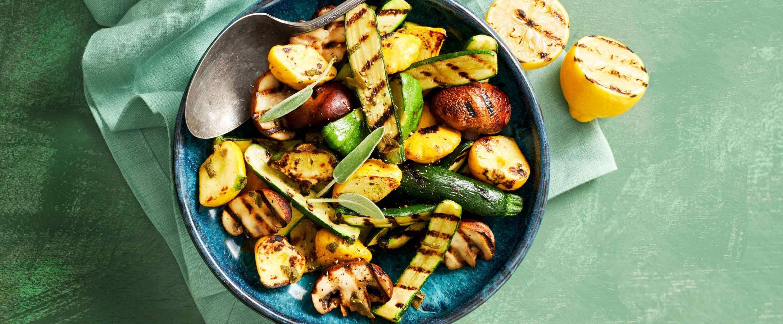 An assortment of grilled zucchini and squash in a blue bowl, with grilled lemon halves off to the side on the table