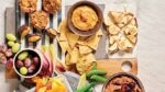 an array of vegan movie night snacks recommended by plant based pros, spread on a striped tablecloth. Snacks include stuffed dates, frozen grapes, snap peas, and banana toast
