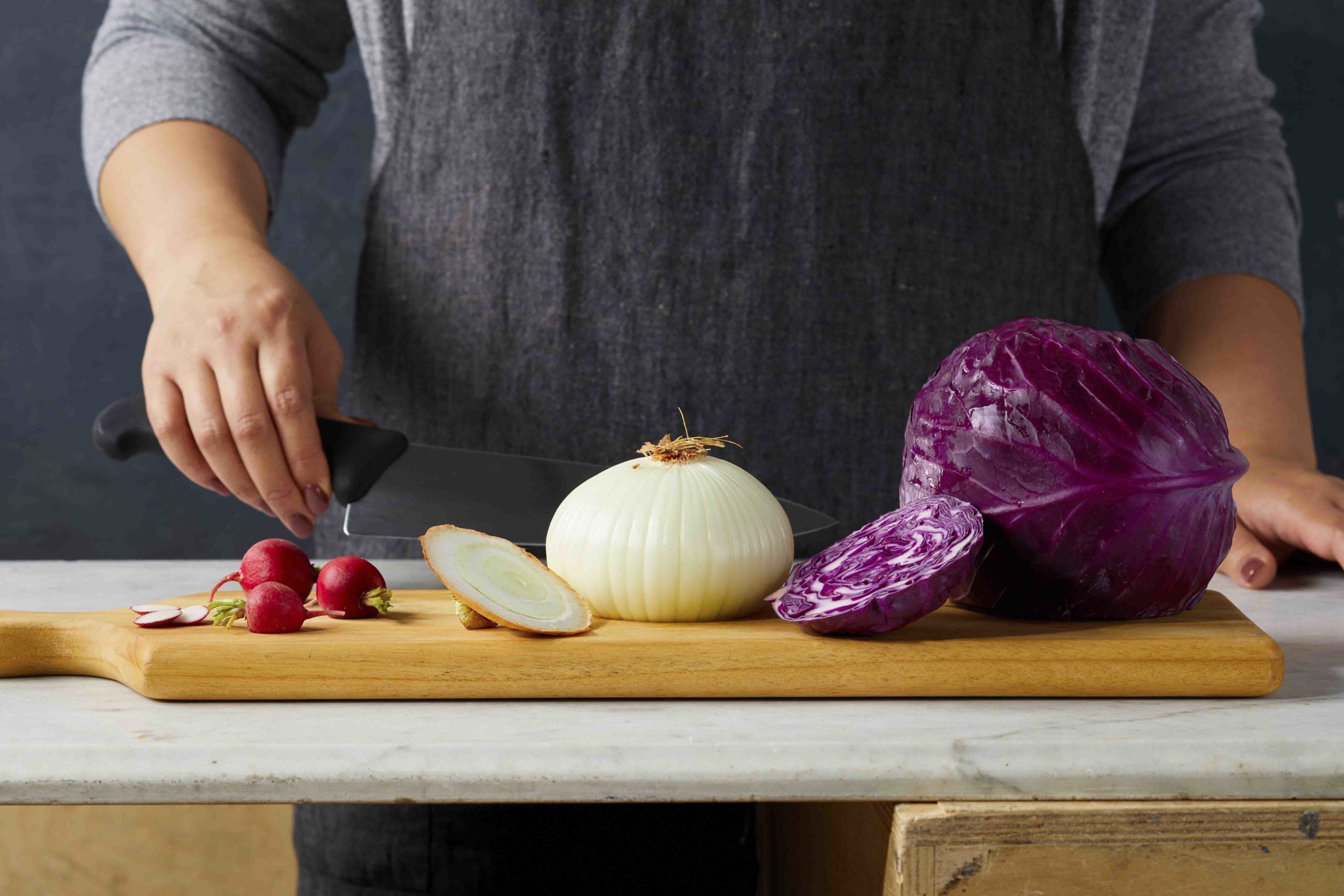 The midsection of a person standing behind a cutting board on a counter; on the cutting board are radishes, onions, cabbage, being cut into blocks to help create stability for slicing and dicing