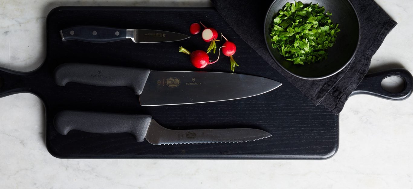 A dark cutting board with three knives laid out parallel across the board, along with a few radishes and a small bowl with finely chopped herbs