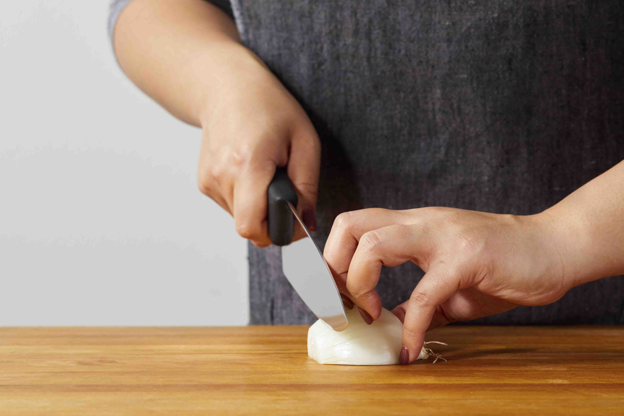 The midsection of a person who stands behind a counter working over a cutting board holding an onion, shaping their hand into a claw to avoid cutting the tips
