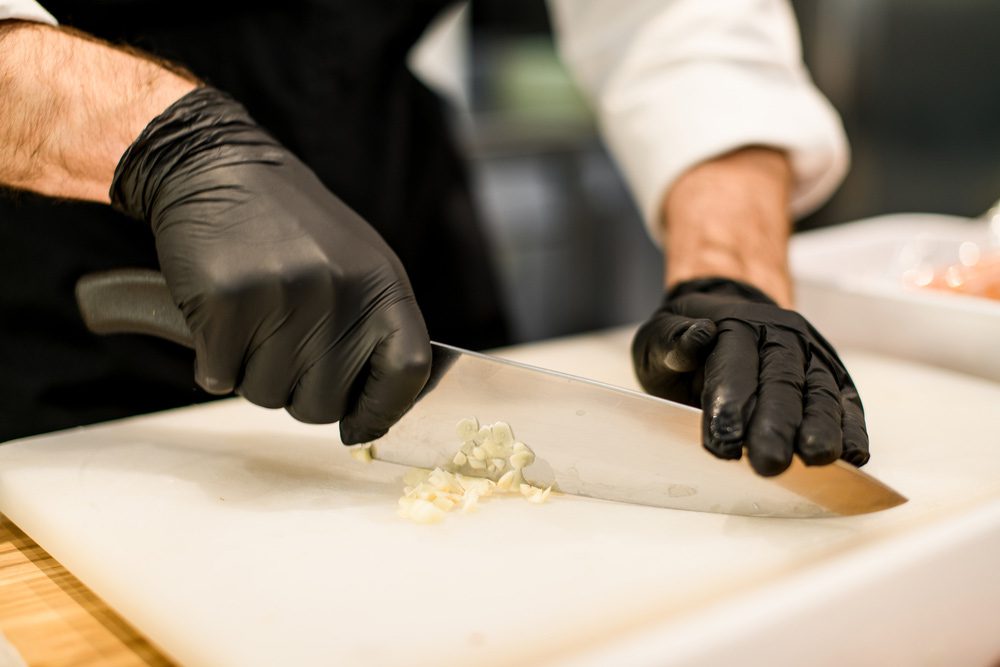 Hands wearing black gloves mincing garlic on a cutting board using a chef's knife