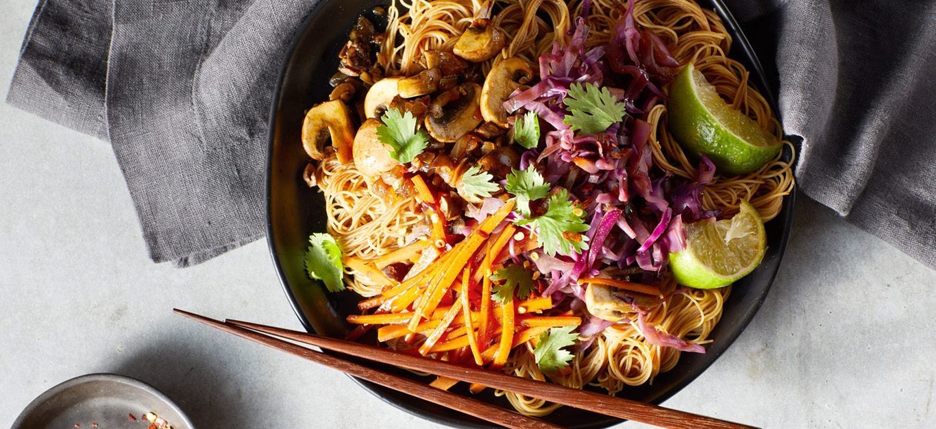Pancit with Vegetables - Plant-based take on a Filipino dish. Thin rice noodles in a bowl with mushrooms and shredded carrots, with lime wedges and chopsticks on the side