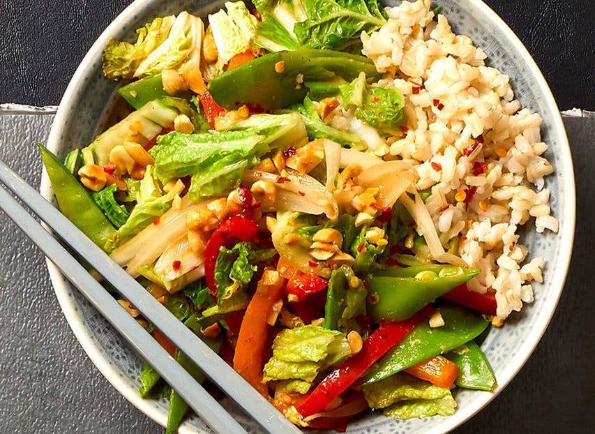 peanut sauce stir-fry - array of shredded cabbage, snap peas, and bell peppers tossed together and served over a bed of brown rice, shown on a gray table next to a glass of sparkling water, with chopsticks on the side