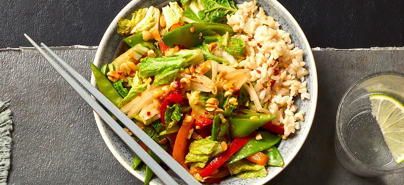 peanut sauce stir-fry - array of shredded cabbage, snap peas, and bell peppers tossed together and served over a bed of brown rice, shown on a gray table next to a glass of sparkling water, with chopsticks on the side