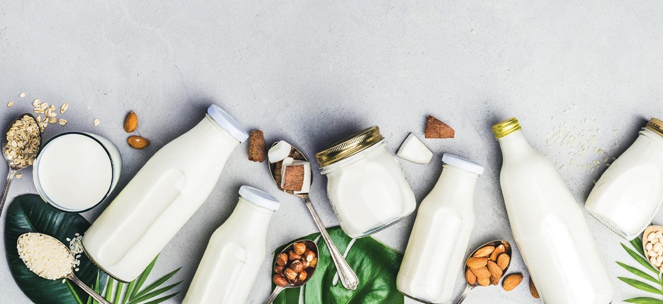 Glass bottles of various types of nondairy plant milks, including oat, soy, and nut, shown laid next to each other, with nuts, soybeans, and leaves in between
