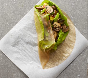 A half-rolled rice paper wrap, with a filling of lettuce, mushrooms, and rice, sitting atop a square of parchment paper