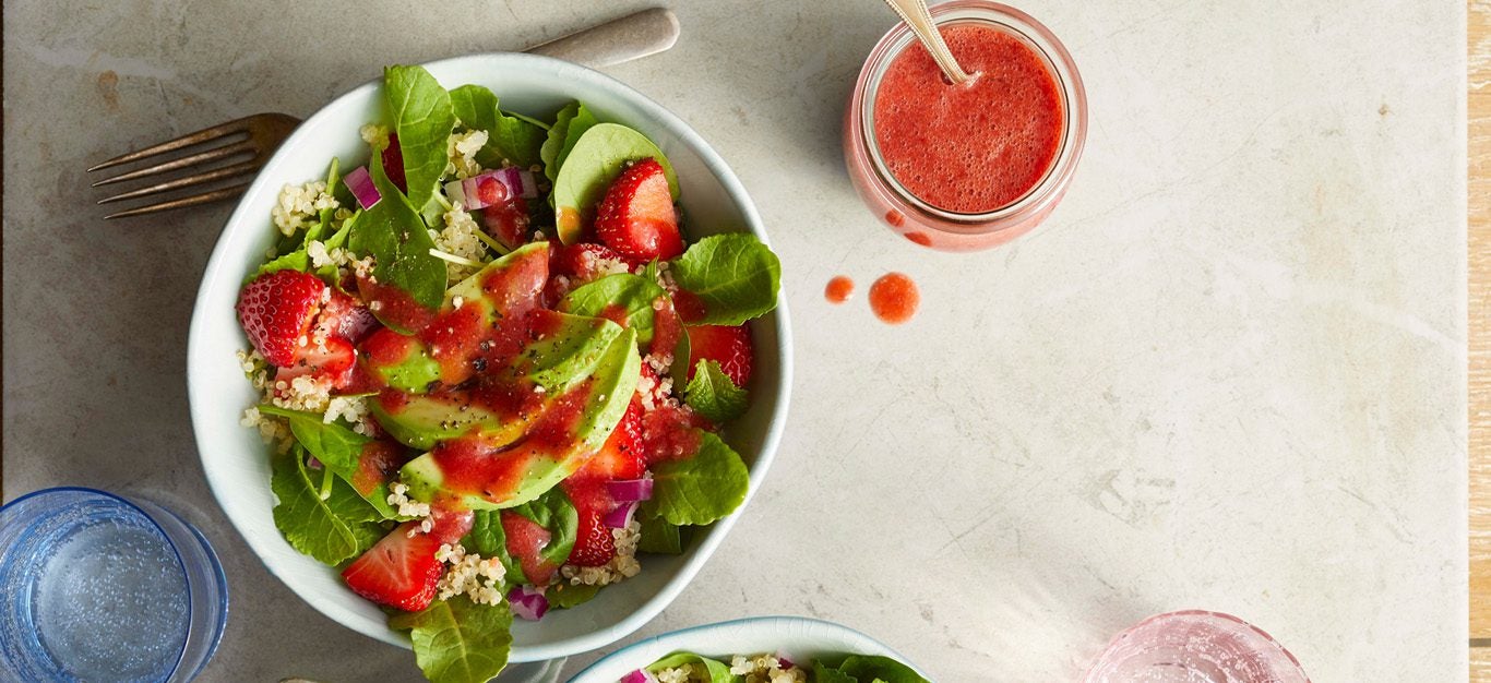 A colorful salad with sliced strawberries, avocado, and greens mixed together and drizzled with a bright red strawberry dressing, with a jar of strawberry dressing on the side