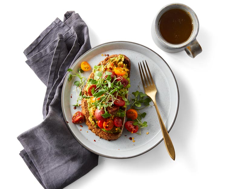 Avocado toast with cherry tomatoes and sprouts on a whitw plate with a gray napkin and cup of coffee in right corner